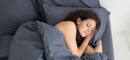 Woman sleeping on a pillow on her side in bed. Bed is made with grey bedding.