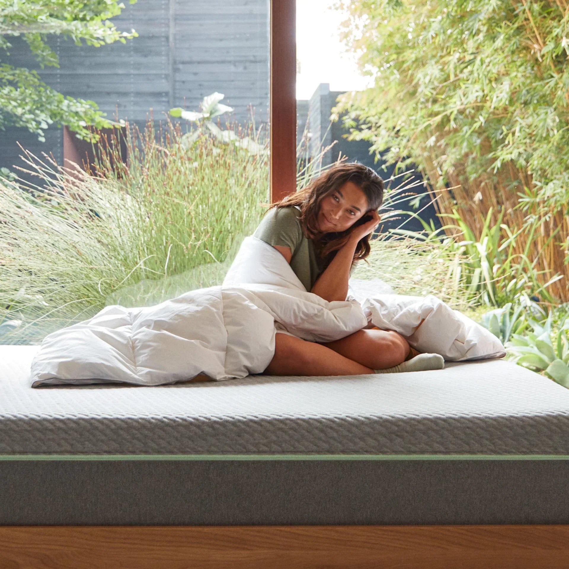 Grey mattress sitting on a light brown bed frame with two white pillows on it with two matching light brown end tables are on each side both contain a lamp on them all in front of a green wall all on top of a white rug