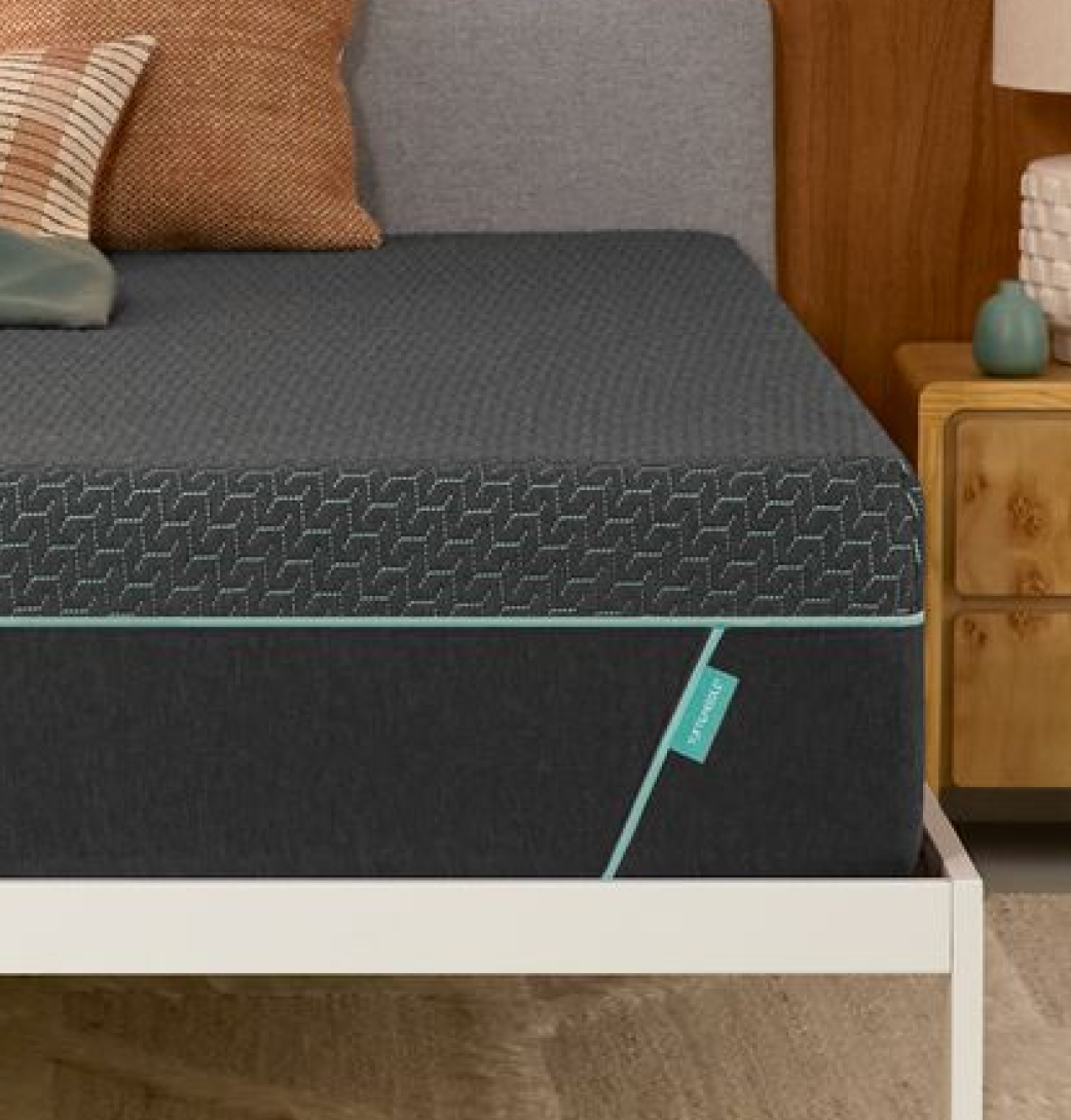 Tuft & Needle Mint Hybrid mattress on a white bed frame with tan and green pillows beside a wooden nightstand.