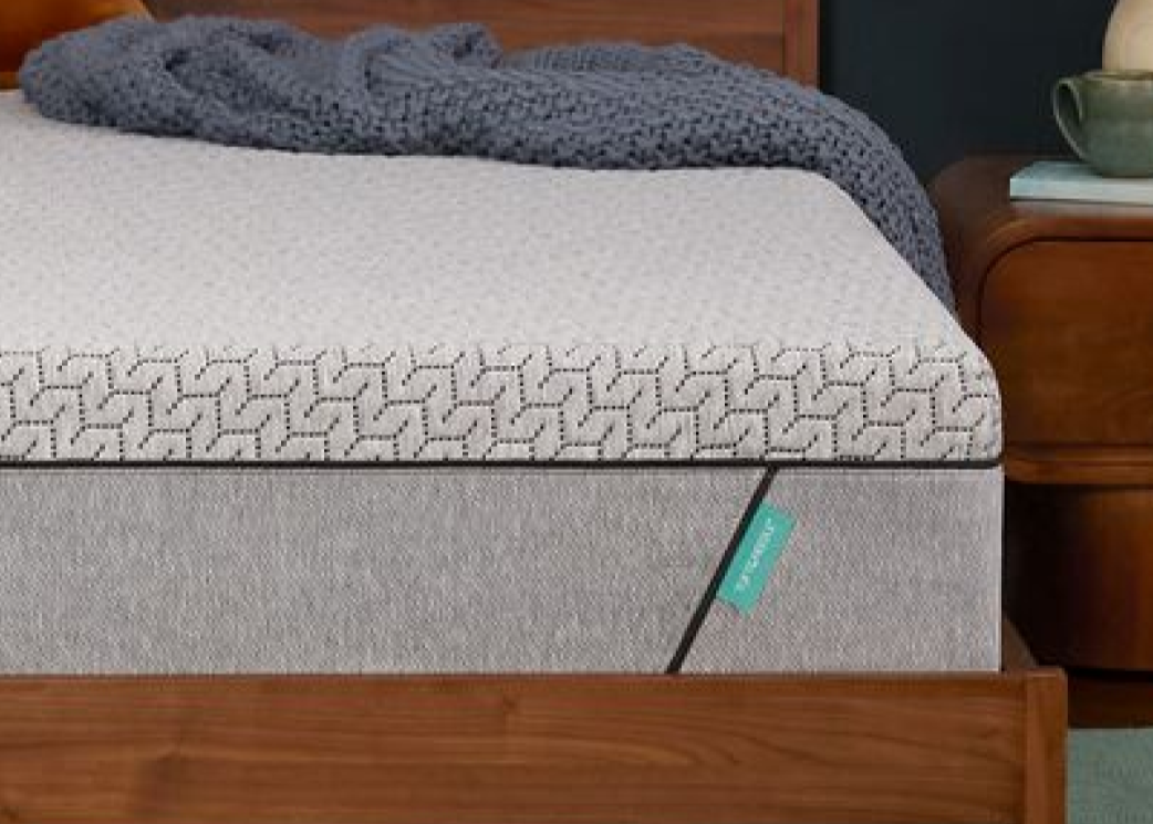 Tuft & Needle Mint mattress on a wooden bed frame with a gray knit blanket and rust-colored pillow.