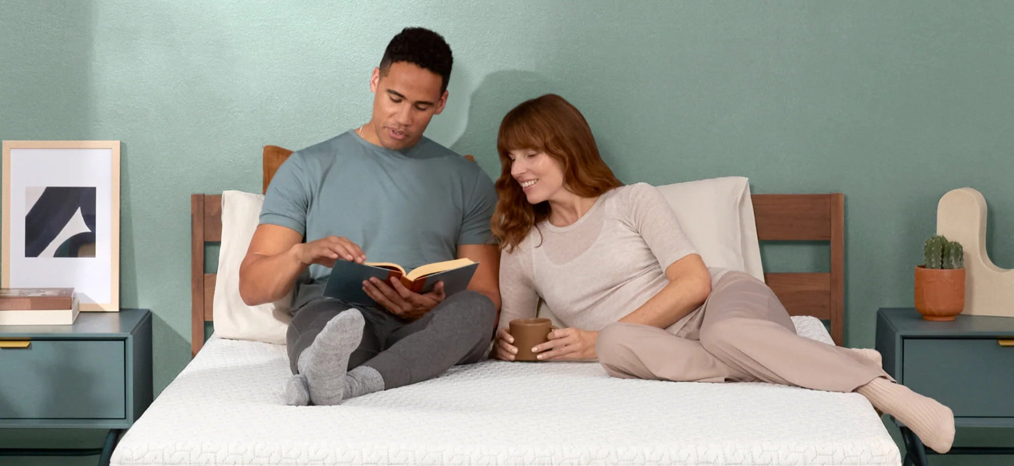 Woman drinking a coffee and a man reading a book on a mattress 